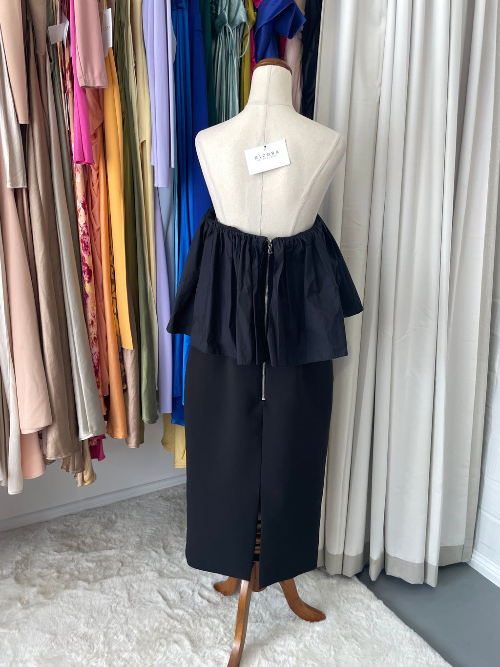 Black dress on a mannequin with a label, placed on a stack of colorful fabric rolls.