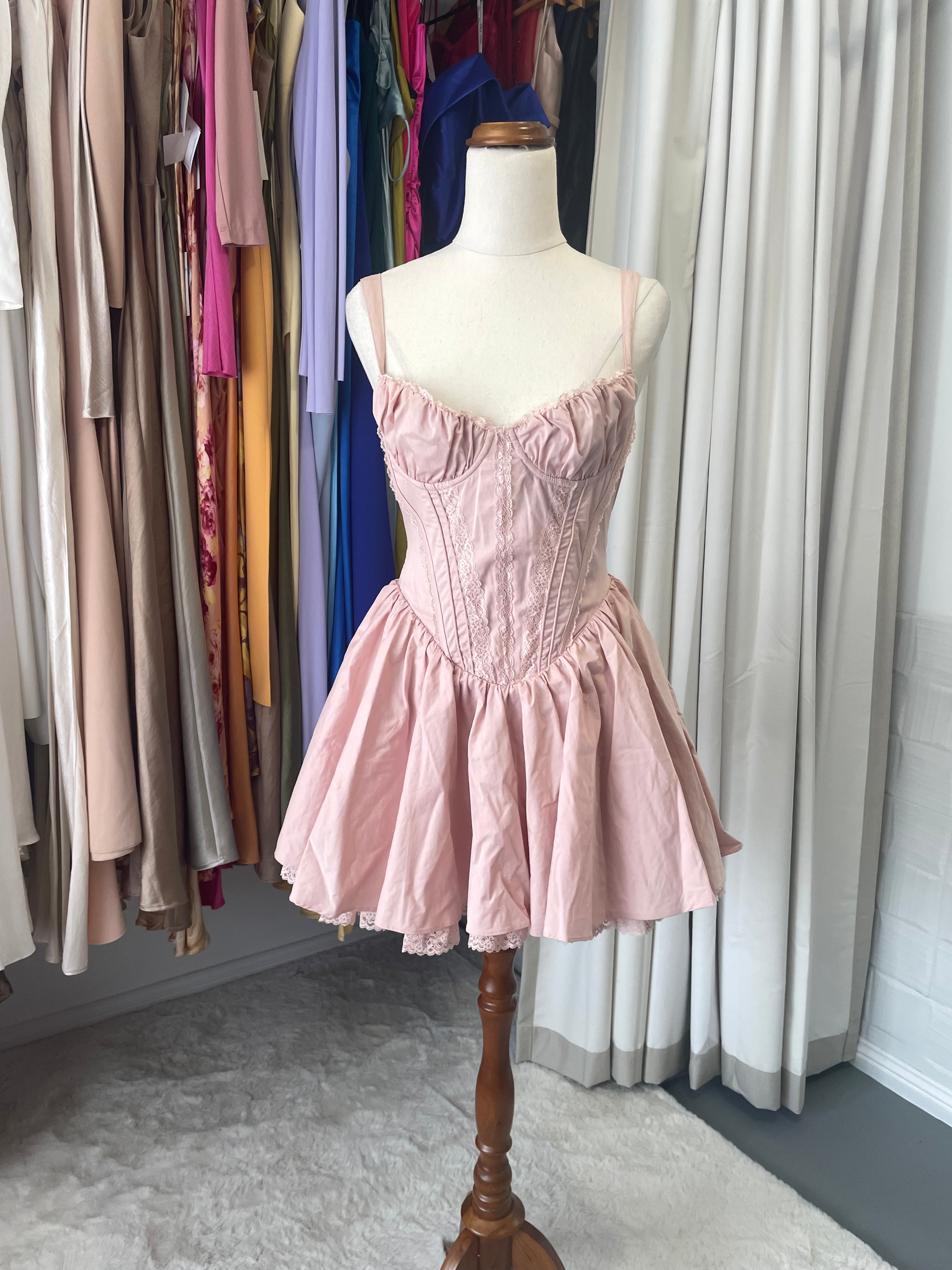 Baby Boo Josie is an elegantly crafted corset dress featuring delicate lace detailing, structured boning for a refined silhouette, and a graceful full skirt. The blush tone and tie bow back create a romantic aesthetic, while underwired cups and full lining ensure comfort and support.