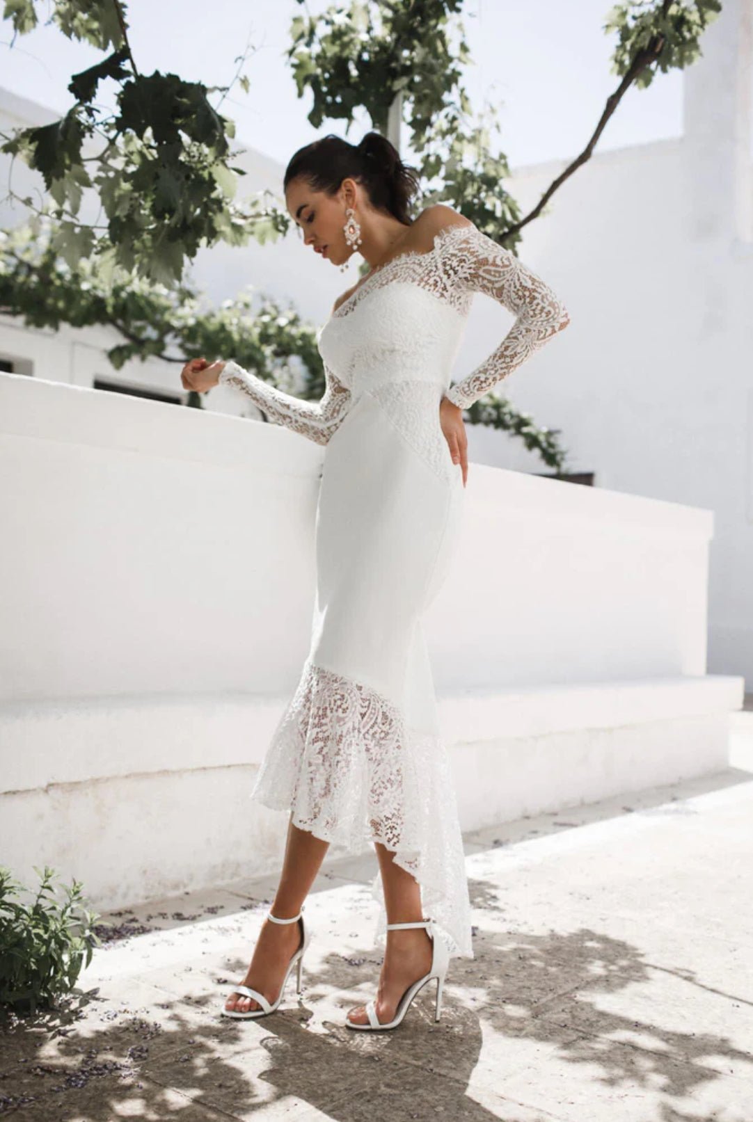 Woman in a white lace wedding reception dress standing outdoors with white walls and greenery.