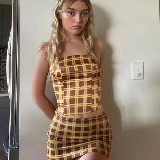 Woman wearing a yellow and brown plaid dress standing against a plain wall.
